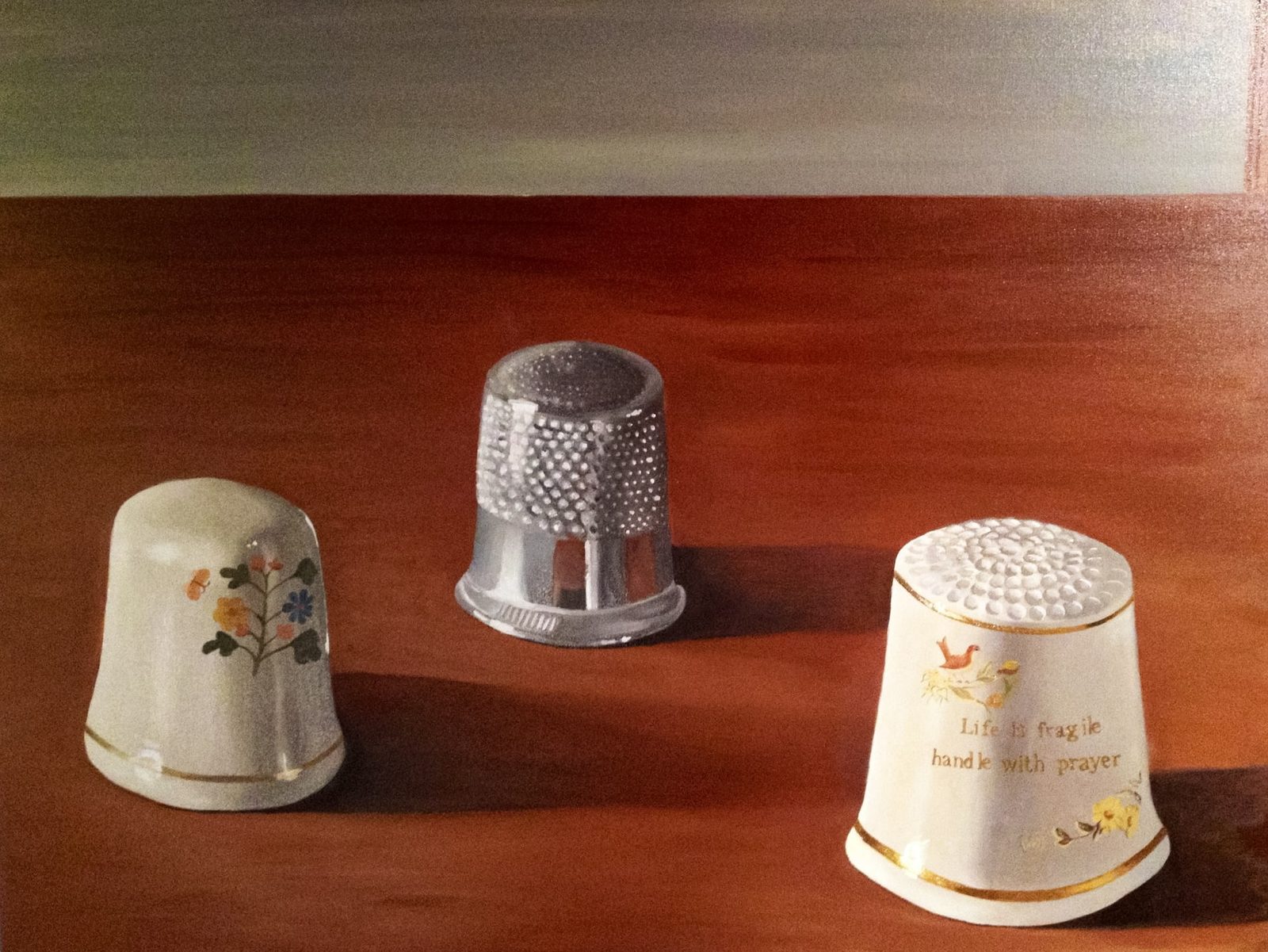 Three thimbles on a wood surface: floral porcelain, silver, and a porcelain thimble printed 'life is fragile, handle with prayer'.