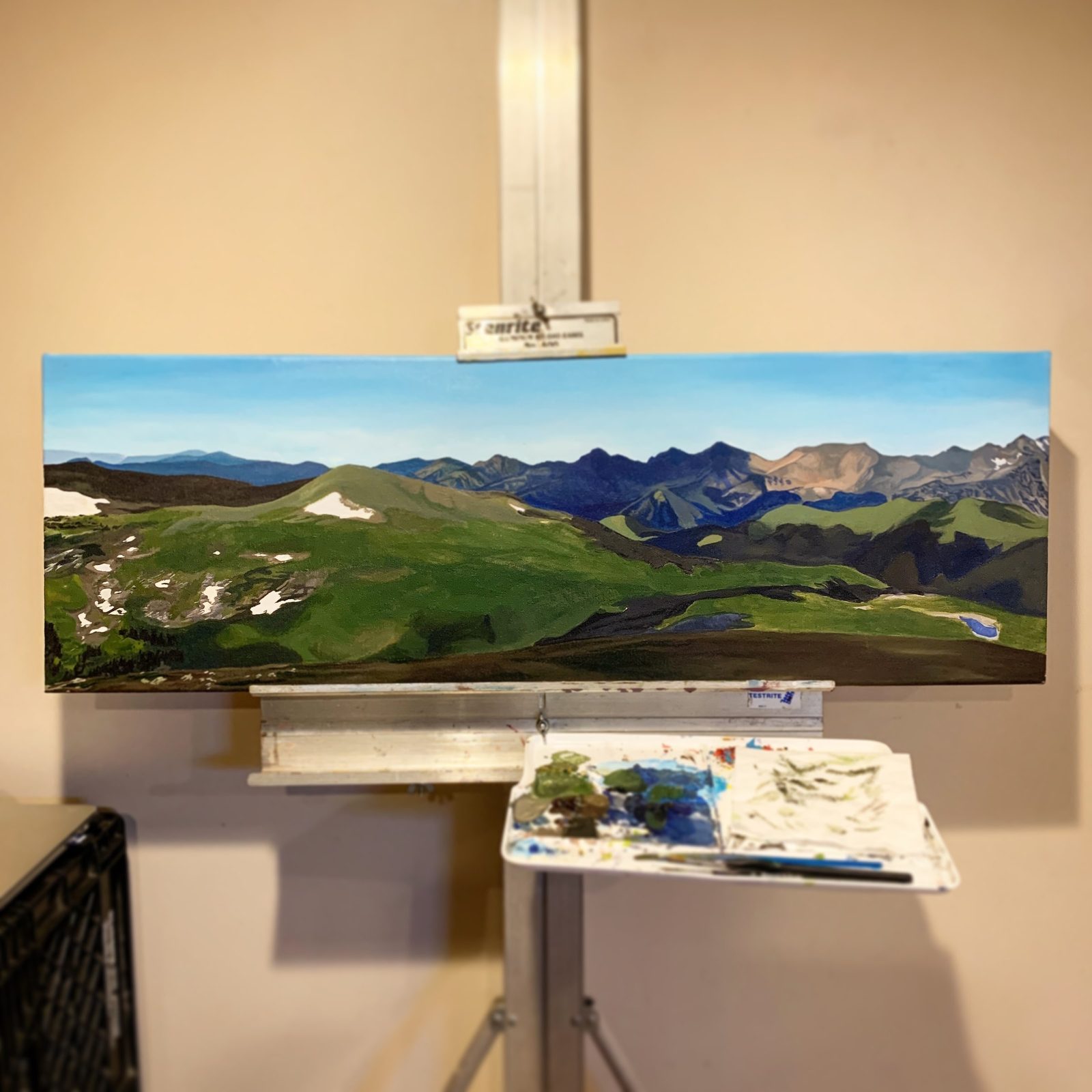 Landscape painting of green mountains with snow patches under a blue sky, displayed on an easel.