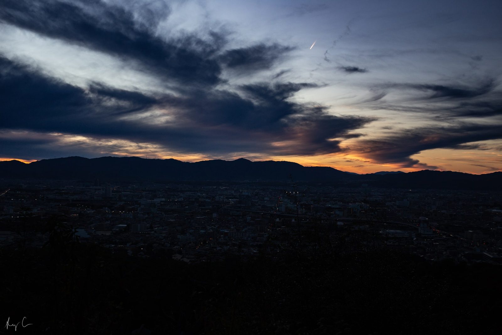 City skyline at dusk beneath dark hills and a streaked orange sunset