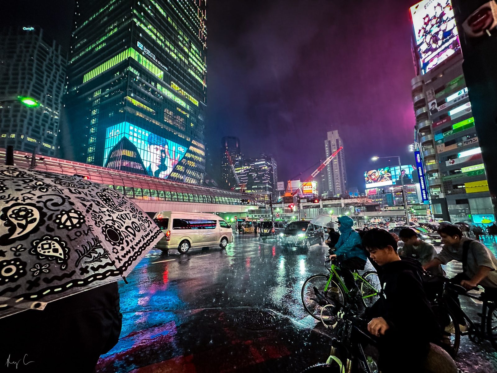 Rainy night city intersection with illuminated signs, an umbrella, and cyclists waiting