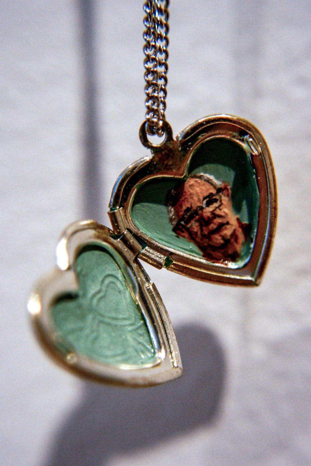 Open gold heart-shaped locket with teal enamel interior, holding a painted portrait of a man wearing glasses.