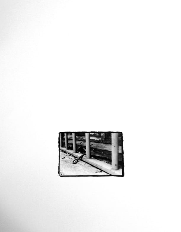 Small black-and-white print, rough-edged on a white ground, showing a coated metal fence with vertical posts and a key hanging near its paved base.