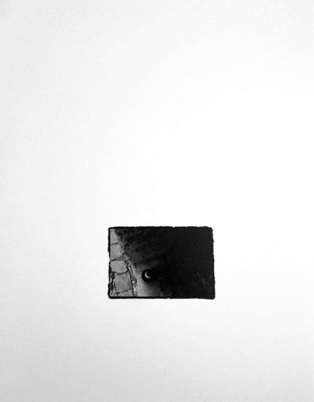 Small black-and-white print, rough-edged on a white ground, showing a dark round object in a cobbled window sill with a small reflective highlight.