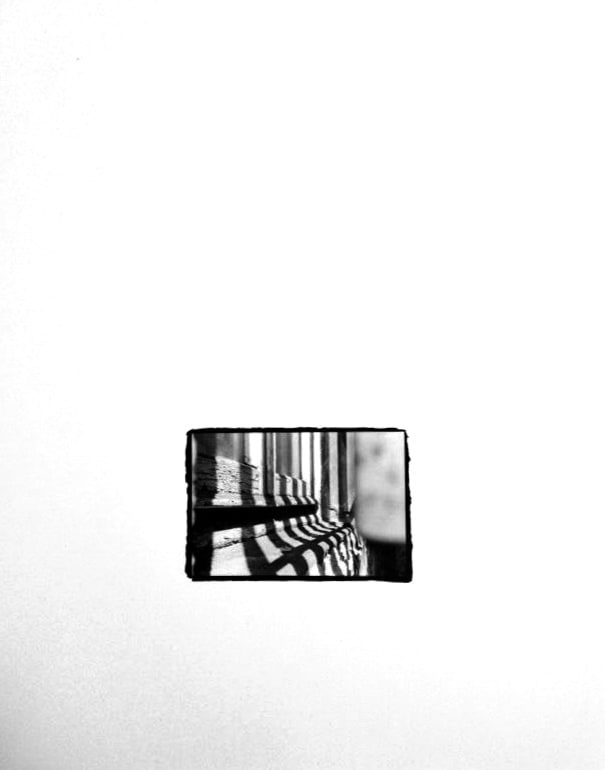 Small black-and-white print, rough-edged on a white ground, showing vertical bars casting long striped shadows across a wooden frame.